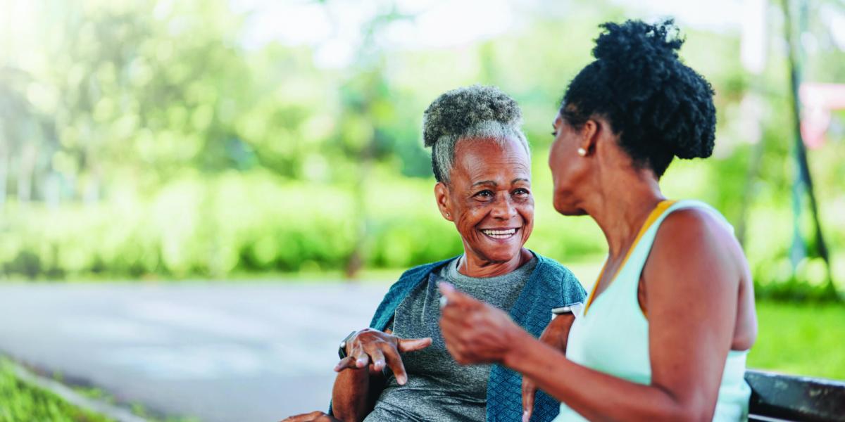 Photograph of two joyful Black women chatting on a bench in a sunny park.